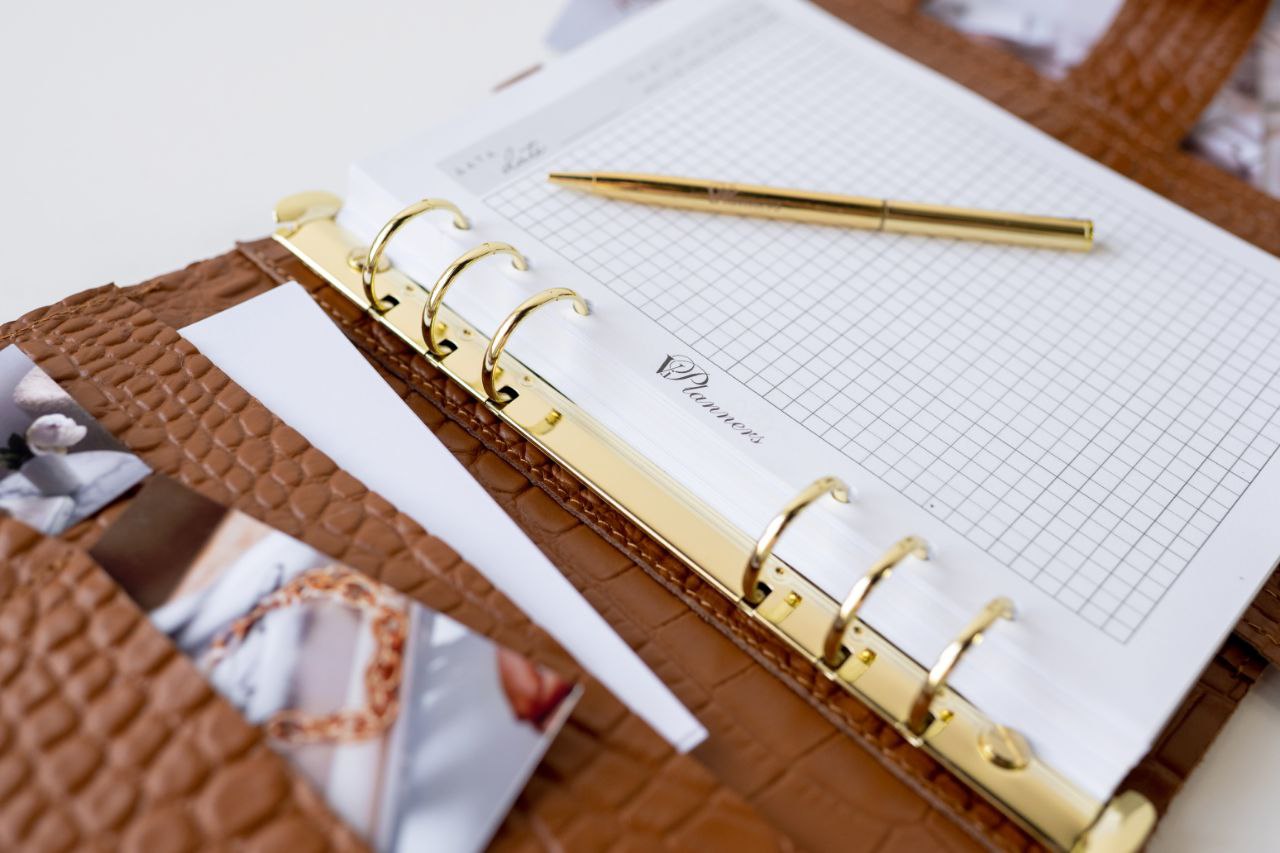 Close-up of a brown leather A5 notebook cover with a gold binder and grid paper, featuring a pen and documents.